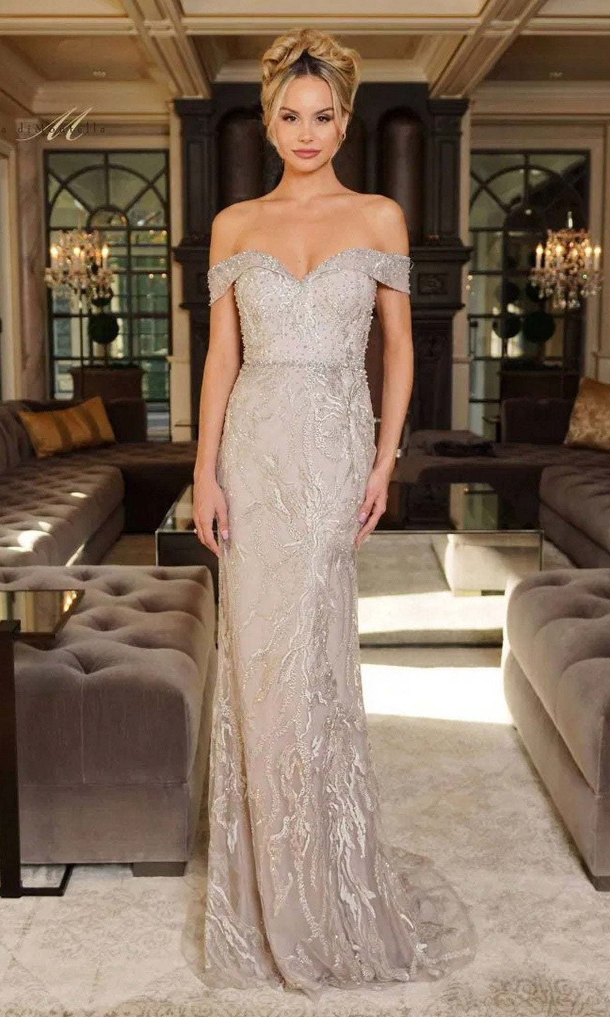 A woman stands in an elegant living room wearing the Rina Di Montella RD2713—a long formal dress in light mauve, adorned with intricate beading and lace embellishments.